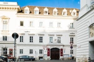 Ein historisches europäisches Gebäude mit cremefarbener Fassade, mehreren Fenstern und einem steilen Ziegeldach bietet die ideale Kulisse für die Hochzeitsplanung. Es verfügt über dekorative Akzente und eine zentrale Holztür. Mehrere geparkte Autos und Verkehrsschilder säumen die Straße unter dem klaren blauen Himmel.