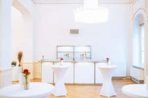 Ein heller, eleganter Raum, perfekt für eine Hochzeit, mit drei hohen Tischen, die mit weißen Tischdecken bedeckt sind und jeweils mit einer kleinen Vase mit roten Blumen geschmückt sind. Im Hintergrund ist ein minimalistischer Barbereich sichtbar. Ein großer Kronleuchter hängt von der Decke und beleuchtet den Holzboden.