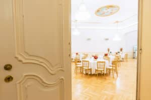 Eine offene Tür gibt den Blick auf einen eleganten Speisesaal frei, der sich perfekt für eine Hochzeit eignet. Die runden Tische sind mit weißen Tischdecken bedeckt und mit roten Blumen geschmückt. Goldene Stühle sind um die Tische herum angeordnet und ein dekoratives Deckenmotiv mit Kronleuchtern wertet die Atmosphäre für Ihren besonderen Tag auf.
