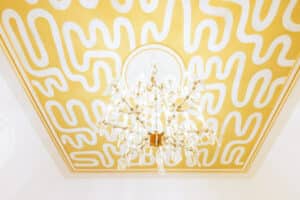 Ein Kristallkronleuchter hängt elegant von einer Decke, die mit einem abstrakten weiß-gelben Muster bemalt ist – ideal für alle, die gerade mitten in der Hochzeitsplanung stecken. Das kühne Design mit seinen Wellenlinien erzeugt einen modernen, künstlerischen Effekt rund um die kunstvolle Leuchte, perfekt, um Hochzeitsträume wahr werden zu lassen.