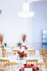 Ein eleganter Speisesaal für eine Hochzeit, mit runden Tischen, die für die Veranstaltung arrangiert wurden. Jeder Tisch ist mit roten Blumendekorationen, weißen Tischdecken und ordentlich arrangiertem Glasgeschirr geschmückt. Ein großer Kristallkronleuchter hängt darüber und verbreitet ein sanftes Licht, das perfekt für Hochzeiten ist.