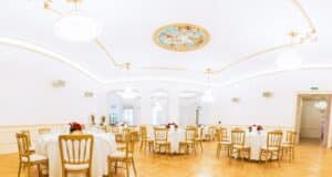 Ein heller, eleganter Speisesaal, der sich perfekt für die Hochzeitsplanung eignet, mit runden Tischen für die Gäste. Jeder Tisch ist mit einer weißen Tischdecke bedeckt und mit roten Blumen geschmückt. Der Raum verfügt über kunstvolle Deckenkunstwerke, Kronleuchter und große Fenster, ergänzt durch den polierten Holzboden.