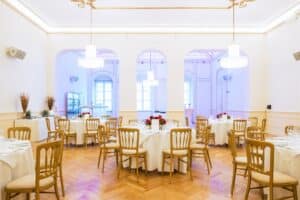 Ein heller, eleganter Bankettsaal, der sich perfekt für eine Hochzeit eignet, mit runden Tischen, die mit roten Blumen geschmückt sind. Der Raum verfügt über hohe Decken, gewölbte Türen, Holzböden und kunstvolle Kronleuchter, die ein anspruchsvolles Ambiente schaffen, das sich ideal für die Hochzeitsplanung eignet.