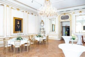 Eleganter Speisesaal mit Parkettboden, verzierten weißen und goldenen Wänden und einem großen Kronleuchter – perfekt für eine Hochzeitsfeier. Mehrere runde Tische sind mit weißen Tischdecken, Blumengestecken und hohen Kerzen gedeckt. An der Wand hängt ein großes Porträt.