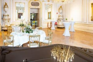 Ein luxuriöser Bankettsaal mit kunstvollen Goldverzierungen und Kronleuchtern, perfekt für Ihre Hochzeit. Runde Tische, elegant gedeckt mit weißen Tischdecken und Blumendekorationen, während hohe Cocktailtische in der Ecke stehen. Ein großer Spiegel reflektiert die Kronleuchter und verleiht Ihrer Hochzeitsplanung einen Hauch von Magie.