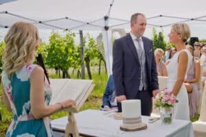 Ein Paar steht lächelnd während einer Hochzeitszeremonie im Freien unter einem weißen Baldachin da. Eine Frau in einem geblümten Kleid führt die Zeremonie durch, im Hintergrund sitzen die Gäste. Ein Blumenstrauß und eine Kerze schmücken den Tisch und sorgen für einen bildschönen Moment ihrer Hochzeitsfeier.