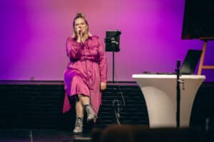 Eine Person in einem langärmeligen rosa Kleid sitzt auf der Bühne, hält ein Mikrofon und singt und schafft so die perfekte Atmosphäre für eine Hochzeitsfeier. Der Hintergrund leuchtet in rosa und violetten Lichtern. Sie trägt eine Brille und Stiefel, und ein Ständer und ein kleiner Tisch in der Nähe vervollständigen die Szene.