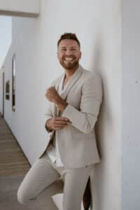 Ein Mann in einem hellbeigen Anzug steht vor einer weißen Wand und lächelt strahlend, als wäre er bereit für seine Hochzeit. Er hat kurze Haare und einen gepflegten Bart. Im Hintergrund ist ein Holzweg zu sehen, der in ein Gebäude führt. Die Szene ist gut beleuchtet und lässt auf einen sonnigen Tag schließen, der perfekt für eine Hochzeit ist.