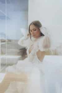 Eine Frau in einem zarten, durchsichtigen weißen Kleid sitzt anmutig da und verkörpert die Eleganz einer Hochzeit, während ihre Hände den Tüll um ihre Schultern zurechtrücken. Der Raum strahlt eine sanfte, ätherische Atmosphäre mit gedämpften Farben und einer Glastrennwand im Hintergrund aus.