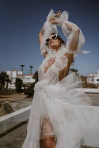 Eine Frau in einem fließenden, durchsichtigen weißen Kleid posiert mit erhobenen Armen vor einem klaren Himmel und verkörpert den Geist einer Hochzeit. Sie trägt eine herzförmige Sonnenbrille und steht auf einem Dach mit weißen Gebäuden und Palmen im Hintergrund. Die Szene strahlt eine luftige, stilvolle Atmosphäre aus, die perfekt für die Hochzeitsplanung ist.