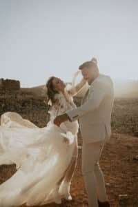 Ein Paar tanzt fröhlich in einer Wüstenlandschaft und feiert seine Hochzeit. Die Frau trägt ein fließendes weißes Kleid und der Mann einen hellen Anzug. Die Sonne wirft einen warmen Schein und schafft eine romantische und verträumte Atmosphäre, die perfekt für einen so intimen Moment der Hochzeit ist.