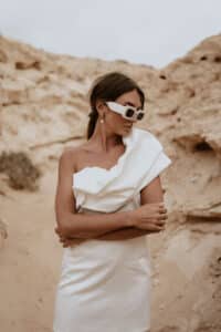Eine Frau in einem eleganten weißen Kleid und mit Sonnenbrille steht in einer sandigen, felsigen Wüstenlandschaft, die an eine einzigartige Hochzeitskulisse erinnert. Sie hat die Arme verschränkt und blickt zur Seite. Dramatische Felsformationen bilden eine beeindruckende Kulisse.