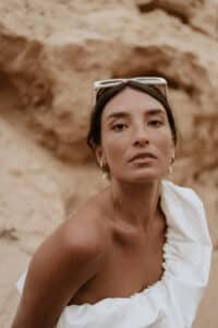 Frau mit dunklem Haar und Creolen, einem weißen One-Shoulder-Top und einer Sonnenbrille mit weißem Rahmen auf dem Kopf steht zwischen Sandsteinen. Sie blickt direkt in die Kamera und strahlt die Eleganz eines Hochzeitsmoments aus.