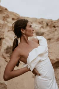 Eine Frau in einem weißen, schulterfreien Kleid steht mit der Hand auf der Hüfte. Ihr Haar ist zurückgebunden und wird durch elegante Ohrringe betont. Vor beigefarbenen Felsformationen posierend, verkörpert sie die heitere Stimmung der Hochzeitsplanung und blickt nachdenklich zur Seite.