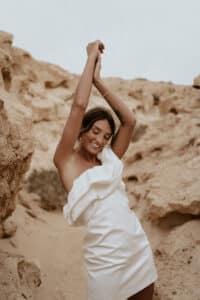 Eine Frau in einem schulterfreien weißen Kleid streckt freudig ihre Arme in die Höhe, während sie in einer felsigen Wüstenlandschaft steht und die Essenz von Hochzeit einfängt. Die natürlichen Beigetöne der Felsen ergänzen ihr Outfit und bilden eine perfekte Kulisse für Heiraten.
