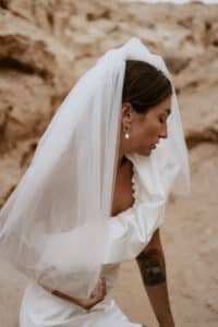Inmitten einer Wüstenlandschaft kniet eine Frau in einem weißen Brautkleid und Schleier anmutig nieder. Ihr Haar ist zurückgebunden, sie trägt goldene Ohrringe und zeigt ein Tattoo auf ihrem Arm. Die Szene vor sandigen Felsformationen fängt die erhabene Ruhe des Hochzeitsglücks ein.