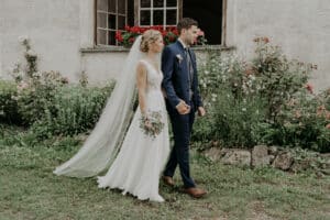 Braut und Bräutigam gehen Hand in Hand im Freien und verkörpern die Essenz einer wunderschönen Hochzeit. Die Braut trägt ein weißes Kleid mit langem Schleier und hält einen Blumenstrauß, während der Bräutigam einen blauen Anzug trägt. Sie stehen vor einem mit Blumen geschmückten Garten und einem rustikalen Gebäude, perfekt für ihre Hochzeitsplanungsträume.