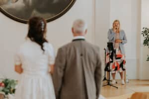 Eine Frau in blauem Blazer und gemustertem Rock spricht auf einer kleinen Bühne in ein Mikrofon und führt das Paar durch ihre Hochzeitsplanung. Im Vordergrund stehen sie mit dem Rücken zur Kamera, ihr zugewandt. Der Raum hat helle Wände und darüber ein großes rundes Gemälde.