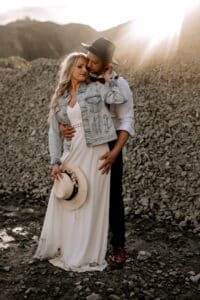 Ein Paar umarmt sich bei Sonnenuntergang in einer felsigen Landschaft und fängt einen romantischen Moment ein, der ihre Hochzeitsträume widerspiegelt. Die Frau in Jeansjacke und weißem Kleid hält einen Hut, während der Mann in weißem Hemd und dunklem Hut hinter ihr steht und sanft ihre Schultern hält, während das Sonnenlicht über sie strömt.