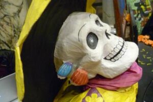 Eine farbenfrohe Skelettfigur zum Tag der Toten mit breitkrempigem Hut und Ohrringen, die an Hochzeitsfeiern erinnern. Die Figur ist wie eine festliche Braut in spe mit heller Kleidung und einem violetten Schal geschmückt und verkörpert sowohl den fröhlichen Geist des Anlasses als auch traditionelle Elemente.