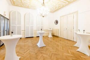 Eleganter Raum mit einem hölzernen Fischgrätboden, hohen gewölbten Türen und einem Kronleuchter. Mehrere Cocktailtische mit weißen Bezügen und kleinen Blumenarrangements sind gleichmäßig verteilt. Perfekt für die Hochzeitsplanung, die weißen Wände mit goldenen Akzenten schaffen eine edle Atmosphäre.