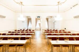 Ein geräumiger, elegant eingerichteter Konferenzraum mit Parkettboden, der sich perfekt für Hochzeitsplanung eignet. Tisch- und Stuhlreihen sind ordentlich nach vorne ausgerichtet, wo große Fenster mit fließenden Vorhängen natürliches Licht hereinlassen. Kronleuchter hängen von der Decke.