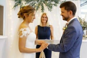 Braut und Bräutigam tauschen bei ihrer Hochzeitszeremonie die Ringe aus. Die Braut trägt ein weißes Kleid mit Spitzenärmeln, während der Bräutigam einen blauen Anzug trägt. Im Hintergrund steht eine Frau, die die Trauung mit Anmut vornimmt. Drapierte Stoffe und Grünpflanzen spiegeln die sorgfältige Hochzeitsplanung in dem wunderschön dekorierten Raum wider.
