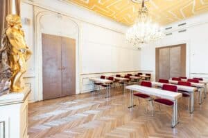 Ein heller, eleganter Raum, perfekt für eine Hochzeit, mit Parkettboden und verzierter Stuckarbeit an der Decke, mit einem großen Kronleuchter. Der Raum hat schmale Tische und rote Stühle in Reihen. In der Ecke neben den doppelten Holztüren steht eine goldene Statue.