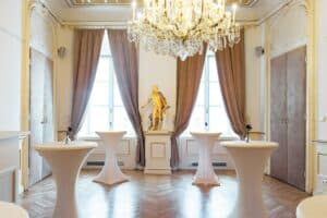 Ein eleganter Raum, der sich ideal für eine elegante Hochzeitsplanung eignet. Er verfügt über einen großen Kronleuchter, drei runde Cocktailtische mit beigen Bezügen und braune Vorhänge, die zwei hohe Fenster einrahmen. In der Mitte an der Rückwand steht auf einem Sockel eine goldene Statue.