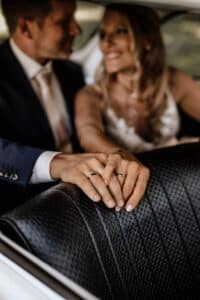 Ein Paar sitzt eng beieinander auf dem Rücksitz eines Autos, ihre Hände sind sanft ineinander verschränkt. Die Frau lächelt den Mann an, beide tragen Eheringe, während sie ihre Hochzeitsplanung besprechen. Der Autositz ist mit einem strukturierten schwarzen Polster versehen, was zur warmen, intimen Atmosphäre beiträgt.