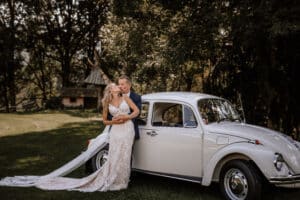 Ein Paar im Hochzeitskleid steht neben einem weißen Oldtimer in einer üppigen, baumbestandenen Umgebung. Der Bräutigam umarmt die Braut, die ein langes Spitzenkleid trägt, das während der sorgfältigen Hochzeitsplanung entworfen wurde. Im Hintergrund ist eine kleine Holzkonstruktion zu sehen.