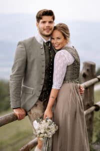 Ein Paar in traditioneller bayerischer Kleidung steht an einem Holzzaun in einer malerischen Umgebung im Freien. Die Frau genießt die Freude ihres Hochzeitstages, hält einen Blumenstrauß in der Hand und lehnt sich liebevoll an den Mann, beide lächeln in die Kamera.
