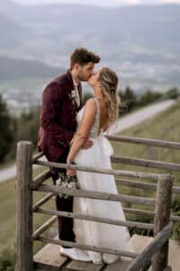Braut und Bräutigam küssen sich auf einem Holzbalkon und fangen so die Essenz ihrer Hochzeit vor einem malerischen Tal ein. Die Braut strahlt in ihrem weißen Kleid, während der Bräutigam einen dunkel gemusterten Anzug trägt. Im Hintergrund schaffen Grün und Berge in der Ferne eine unvergessliche Hochzeitskulisse unter einem bewölkten Himmel.