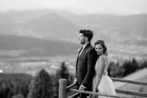 Braut und Bräutigam stehen auf einem Holzbalkon und genießen die Hochzeit mit Blick auf eine weite Berglandschaft. Der Bräutigam blickt nach vorne und hat einen nachdenklichen Blick, während die Braut lächelnd an ihn gelehnt ist. Die Szene ist in Schwarzweiß festgehalten.