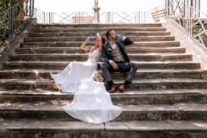 Ein Paar im Hochzeitskleid sitzt auf Steinstufen und verkörpert die Freude am Heiraten. Die Braut lehnt sich an den Bräutigam, während beide aus Wasserflaschen nippen. Sonnenlicht erhellt ihren entspannten und verspielten Moment, eingerahmt von einem Metallgeländer – eine perfekte Pause inmitten des Trubels der Hochzeitsplanung.