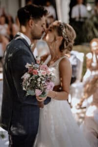 Ein Paar im Hochzeitskleid küsst sich; die Braut hält einen Strauß rosa und weißer Blumen in den Händen und umarmt ihren Partner eng. Die Gäste verfolgen die Zeremonie in dieser idyllischen Hochzeitskulisse im Freien, wo die Liebe unter freiem Himmel erblüht.