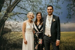 Drei Menschen stehen zusammen vor einer malerischen Kulisse mit einem Baum und einem Feld. Eine Braut in Weiß, ein Mann im Anzug und eine Frau in einem geblümten Kleid lächeln in die Kamera und fangen die freudige Atmosphäre ihrer Hochzeit ein.
