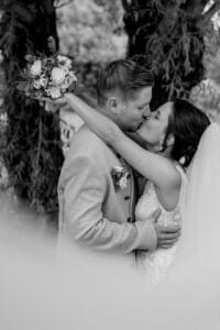 Braut und Bräutigam küssen sich leidenschaftlich im Freien und verkörpern damit die Schönheit ihrer Hochzeit. Die Braut hält einen Blumenstrauß und umarmt ihren Bräutigam in einem hellen Anzug. Sie stehen vor einem Baum, ihr Schleier fällt sanft um sie herum und fängt die Essenz von Liebe und Heirat ein.
