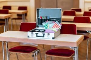 Ein offener Metallkasten mit bunten Markern, runden Aufklebern und Notizkarten steht auf einem Tisch in einem Klassenzimmer mit rot gepolsterten Stühlen und Holztischen und erinnert an die lebhaften Details, die man oft bei einer sorgfältigen Hochzeitsplanung sieht.