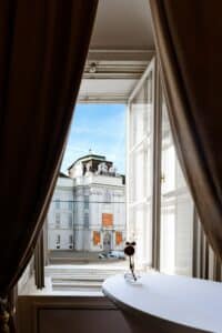 Der Blick aus einem offenen Fenster mit dunklen Vorhängen, die die Szene einrahmen, offenbart ein historisches Gebäude mit klassischer Architektur und Statuen, das an einen märchenhaften Hochzeitsort erinnert. Auf einem kleinen weißen Tisch, der sich ideal für die Hochzeitsplanung eignet, steht eine Vase mit frischen Blumen vor dem Fenster.