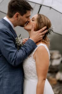 Ein Paar küsst sich romantisch unter einem transparenten Regenschirm an seinem Hochzeitstag. Der Mann trägt einen blauen Anzug, während die Frau ein weißes Spitzenhochzeitskleid mit Blumen im Haar trägt. Der sanft verschwommene Hintergrund lässt den sanften Regen erahnen und sorgt für einen bezaubernden Moment während ihrer besonderen Hochzeitsfeier.