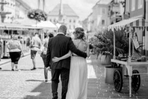 Ein Schwarzweißfoto zeigt ein Paar, das seine Zukunft plant, während es eine Kopfsteinpflasterstraße entlang geht. Der Mann im Anzug und die Frau mit einem Blumenstrauß spiegeln die Essenz von „heiraten“ wider. Sie liegen sich umschlungen, im Hintergrund geschäftige Marktstände.