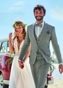 Braut und Bräutigam lächeln, während sie Händchen halten und an einem Strand neben einem rot-weißen Oldtimer spazieren. Die Freude des Heiratens liegt in der Luft. Die Braut trägt ein weißes Kleid und einen Blumenkranz, während der Bräutigam einen grauen Anzug mit Fliege trägt. Ein klarer blauer Himmel bildet die perfekte Kulisse für sie.