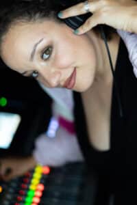 Eine Frau mit Kopfhörern und Ehering blickt mit einem leichten Lächeln in die Kamera. Sie steht vor einem Audiomischpult mit bunten Knöpfen, was darauf schließen lässt, dass sie an der Planung des perfekten Hochzeits-Soundtracks beteiligt ist. Der Hintergrund ist leicht unscharf.