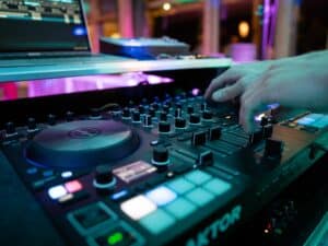 Nahaufnahme der Hände eines DJs, der Knöpfe und Schieberegler an einem digitalen Mischpult einstellt und so den perfekten Ton für eine Hochzeitsfeier einstellt. Das Setup umfasst einen Laptop und einen Traktor-Controller, und bunte Lichter sorgen für eine energiegeladene Atmosphäre, die ideal für die Hochzeitsplanung ist.