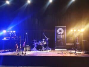 Eine schwach beleuchtete Bühne mit blauen und orangefarbenen Lichtern verfügt über ein Schlagzeug, Keyboards, Gitarren und Mikrofone, perfekt für eine unvergessliche Hochzeit. Ein Banner mit dem Text „CoffeeBreak Jukebox“ steht rechts vor einem schwarzen Vorhang im Hintergrund.