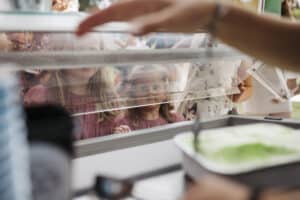Drei Kinder blicken gespannt durch eine Glasvitrine auf eine Auswahl an Eis in einem Geschäft. Ein Kind ist im Fokus und schaut direkt auf das Eis, während zwei andere neben ihm stehen. Der Arm einer Person ist sichtbar, bereit, Leckereien zu servieren, die sich perfekt für jeden freudigen Anlass wie eine Hochzeitsfeier eignen.