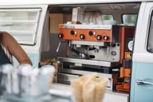 Eine Retro-Kaffeemaschine in einem Oldtimer-Van, perfekt, um jeder Hochzeitsplanung Charme zu verleihen. Kaffeetassen und ein Milchkarton stehen darauf, links ist teilweise eine Person zu sehen. Im Vordergrund sind Waffeltüten auf einer Theke zu sehen.