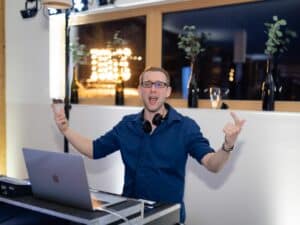 Eine Person in blauem Hemd und Brille steht mit einem Laptop und Kopfhörern um den Hals hinter einem DJ-Setup und gestikuliert enthusiastisch in Richtung einer Hochzeit. Im Hintergrund sind Pflanzen und dekorative Lichter zu sehen, die zur festlichen Atmosphäre beitragen.