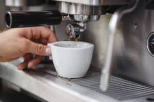 Eine Person hält eine weiße Tasse unter eine glänzende silberne Kaffeemaschine und fängt Espressotropfen auf, als ob sie eine elegante Hochzeit vorbereiten würde. Die Szene zeigt das Ritual der Zubereitung des perfekten Gebräus, ähnlich der Orchestrierung einer nahtlosen Hochzeitsplanung.
